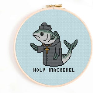 May include: Cross-stitch embroidery in a wooden hoop features a pixelated fish wearing a priest's outfit, including a hat and a cross necklace. The text "HOLY MACKEREL" is below the fish on a light blue background.
