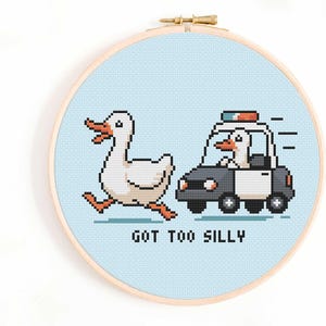 Op de afbeelding: Een borduurring met een gepixeld ontwerp van een witte eend die wegrent van een politieauto die door een eend wordt bestuurd. De tekst "GOT TOO SILLY" staat onder de auto. De achtergrond is lichtblauw.
