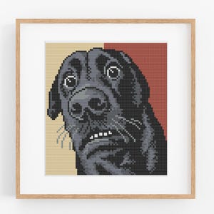 May include: A framed pixel art cross stitch pattern featuring a close-up of a black dog with wide, expressive eyes and bared teeth. The dog is set against a split background of light beige and dark red, all encased in a light wood frame.