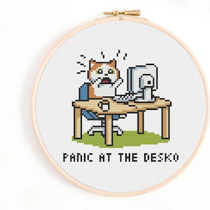 Könnte beinhalten: Ein Kreuzstichmuster mit einer Cartoon-Katze, die an einem Schreibtisch mit einem Computer sitzt und überrascht aussieht. Der Text "Panic at the Desko" befindet sich unter dem Bild.