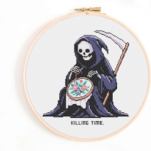 May include: A circular wooden embroidery hoop containing a cross-stitch pattern of a skeleton in a dark purple hooded robe holding a scythe and stitching a floral design. The text KILLING TIME. is stitched in black pixelated letters below the figure.