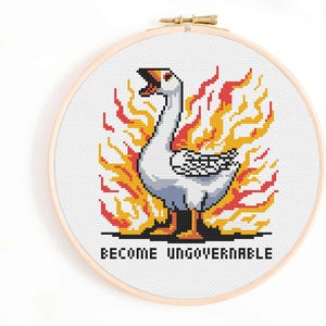 Könnte beinhalten: Kreuzstich-Stickerei in einem Holzrahmen zeigt eine pixelige weiße Gans in Flammen. Die Gans hat einen offenen Schnabel, umgeben von orangefarbenen und gelben Flammen. Der Text "BECOME UNGOVERNABLE" ist darunter gestickt.