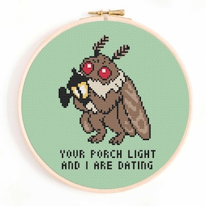 May include: A circular wooden embroidery hoop containing a cross-stitch design of a brown moth with red eyes holding a black lantern. The background is light green fabric. Below the moth, the text reads: YOUR PORCH LIGHT AND I ARE DATING.