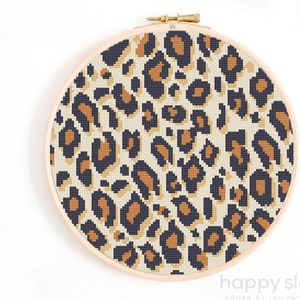 May include: A cross stitch pattern of a leopard print design in a round embroidery hoop. The pattern features black, brown, and beige spots on a cream background.