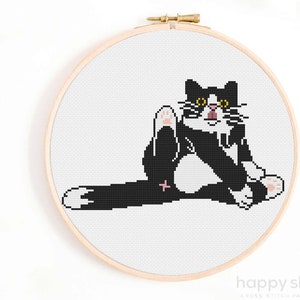 Könnte beinhalten: Kreuzstichmuster einer schwarz-weißen Katze mit gelben Augen, die mit einem hochgehobenen und ausgestreckten Bein sitzt. Die Katze schaut direkt auf den Betrachter.