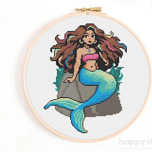 May include: Cross stitch pattern of a mermaid with long brown hair, wearing a pink top and a blue tail, sitting on a rock with seaweed.
