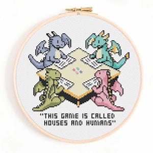 May include: A cross-stitch embroidery in a wooden hoop shows four pixelated dragons around a table. The dragons are blue, green, teal, and pink. The text "THIS GAME IS CALLED HOUSES AND HUMANS" is below the table.