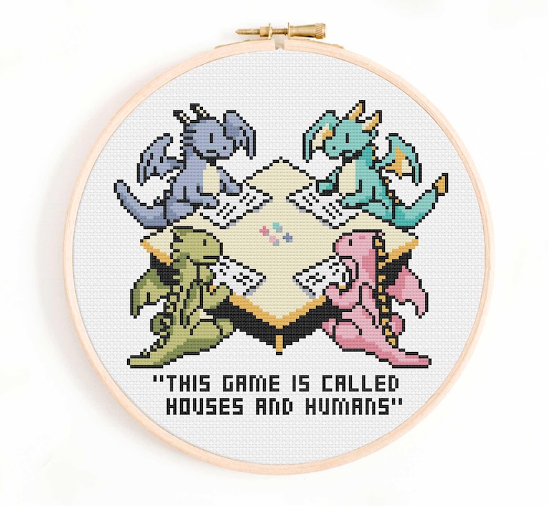 May include: A cross-stitch embroidery in a wooden hoop shows four pixelated dragons around a table. The dragons are blue, green, teal, and pink. The text "THIS GAME IS CALLED HOUSES AND HUMANS" is below the table.