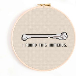 Puede incluir: Un aro de bordado de punto de cruz blanco con una imagen pixelada de un hueso y el texto "Encontré este húmero."