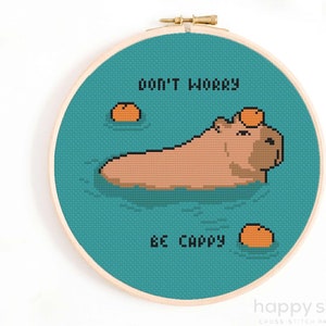 May include: Cross stitch pattern featuring a capybara swimming in turquoise water with the text "Don't worry, be capy" and three orange fruit on top of the capybara and in the water.