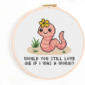 Puede incluir: Bordado de punto de cruz en un aro de madera con un gusano rosa sonriente con una flor amarilla. El texto dice: "WOULD YOU STILL LOVE ME IF I WAS A WORM?" El diseño también incluye hierba verde y tierra marrón.