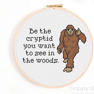 May include: Cross-stitch hoop with the text "Be the cryptid you want to see in the woods." A pixelated brown Bigfoot figure gives a thumbs up. The hoop is set in a wooden frame.