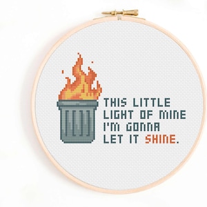 Könnte beinhalten: Kreuzstich-Stickerei in einem Holzreifen. Das Design zeigt einen pixeligen Mülleimer mit Flammen und dem Text "THIS LITTLE LIGHT OF MINE I'M GONNA LET IT SHINE." Der Text und die Flammen sind in Orange- und Rottönen gehalten.