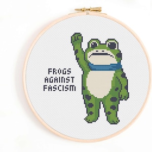 Può includere: Ricamo a punto croce in un telaio di legno con una rana verde a pois neri, con una sciarpa blu e un pugno alzato. Il testo "FROGS AGAINST FASCISM" è sotto. Lo sfondo è bianco.