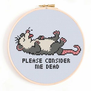 May include: A circular wooden embroidery hoop containing a cross-stitch pattern of a grey and white opossum lying on its back with its tongue out. Below the opossum, the text reads, PLEASE CONSIDER ME DEAD, in black pixelated lettering.