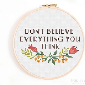 Puede incluir: Bordado de punto de cruz en un aro de madera. El diseño presenta el texto "DON'T BELIEVE EVERYTHING YOU THINK" en letras de bloque marrones, con acentos florales en rojo, verde y naranja. Las palabras están centradas en un fondo blanco.