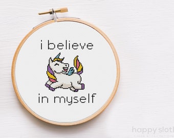Puede incluir: Un bastidor de bordado de punto de cruz con un unicornio blanco con melena, cuerno y alas coloridas. El texto "I believe in myself" está bordado encima y debajo del unicornio. El bastidor de madera tiene un tornillo de latón.