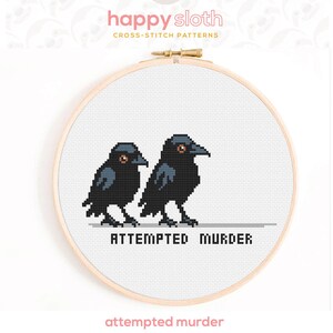 Puede incluir: Patr&oacute;n de punto de cruz con dos cuervos negros con ojos naranjas, uno al lado del otro. La frase "ATTEMPTED MURDER" est&aacute; bordada debajo de las aves. El dise&ntilde;o est&aacute; en un bastidor de bordado de madera.