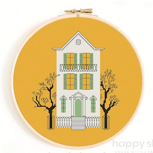 Könnte beinhalten: Kreuzstichmuster eines weißen Hauses mit grünen Fensterläden und einer grünen Tür, vor gelbem Hintergrund. Zwei schwarze Bäume flankieren das Haus. Das Design ist in einem hölzernen Stickrahmen. Der Text "happy sloth" ist sichtbar.