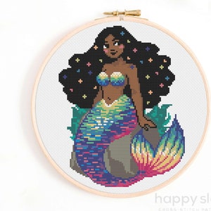 May include: Cross stitch pattern of a mermaid with a rainbow tail and black hair with colorful stars. The mermaid is sitting on a rock with seaweed.