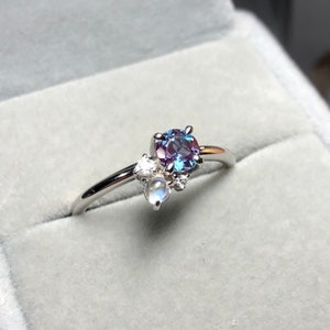 Könnte beinhalten: Ein silberner Ring mit einem blauen und violetten Edelstein und zwei kleineren weißen Edelsteinen. Der Ring befindet sich in einer grauen Samtbox.