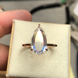 May include: A rose gold ring featuring a large, clear, faceted gemstone. The gemstone is cut in a unique, elongated shape with a pointed top. The ring's band is a simple, thin design, and the gemstone is held in place by four prongs.