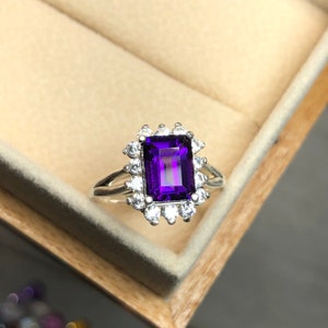 Peut inclure: Une bague en argent avec une grande améthyste rectangulaire violette entourée de petites pierres précieuses claires.