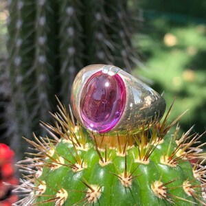 May include: A silver ring with a large, round, pink gemstone set in the center. The ring is sitting on a green cactus.