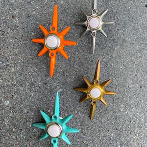 May include: Four starburst-shaped decorative objects in orange, silver, gold, and teal. Each starburst has a white center and pointed rays. These retro-style ornaments are likely for home decor.