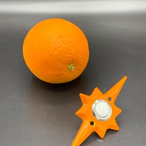 May include: An orange starburst-shaped brooch with a silver-colored center button and two small holes. A whole orange is next to the brooch. The brooch is a bright orange color.