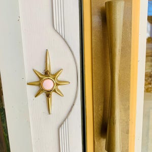 Könnte beinhalten: Eine weiße Tür mit einem goldenen Türgriff und einer goldenen sternförmigen Türklingel mit einem rosa Knopf.