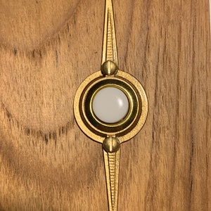 May include: A gold-colored, decorative door knocker with a central white circular element. The design features a circular center with a white button and two elongated, pointed elements extending from the top and bottom. The knocker is set against a wooden background.