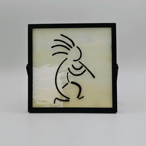 May include: A square, black-framed decorative piece featuring a black line drawing of the Kokopelli figure playing a flute. The background is a light, creamy color, and the frame has a stand on the back.