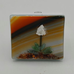 May include: A square glass art piece with a white flower design. The glass has orange, brown, and yellow layers, with a white top. The flower has a white bloom, green leaves, and sits on a bed of brown and orange granules.