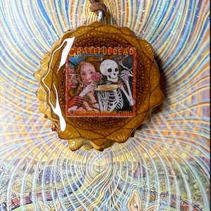 Puede incluir: Un colgante redondo con un borde marrón texturizado y un cordón de cuero marrón. El colgante presenta una imagen colorida con las palabras "Grateful Dead" y un esqueleto sosteniendo una rosa. El fondo tiene un diseño psicodélico.