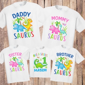 May include: Set of five white t-shirts with dinosaur graphics and text. The shirts say "Daddy Saurus", "Mommy Saurus", "Sister Saurus", "Brother Saurus", and "One-a-Saurus 1 Mason".