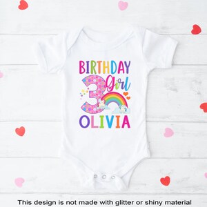 May include: White onesie with a colorful birthday girl design. The number 3 is pink with white polka dots and surrounded by stars. A rainbow with clouds and a unicorn is above the number. The text "Birthday Girl" is in rainbow colors and the name "Olivia" is in rainbow colors below the number.