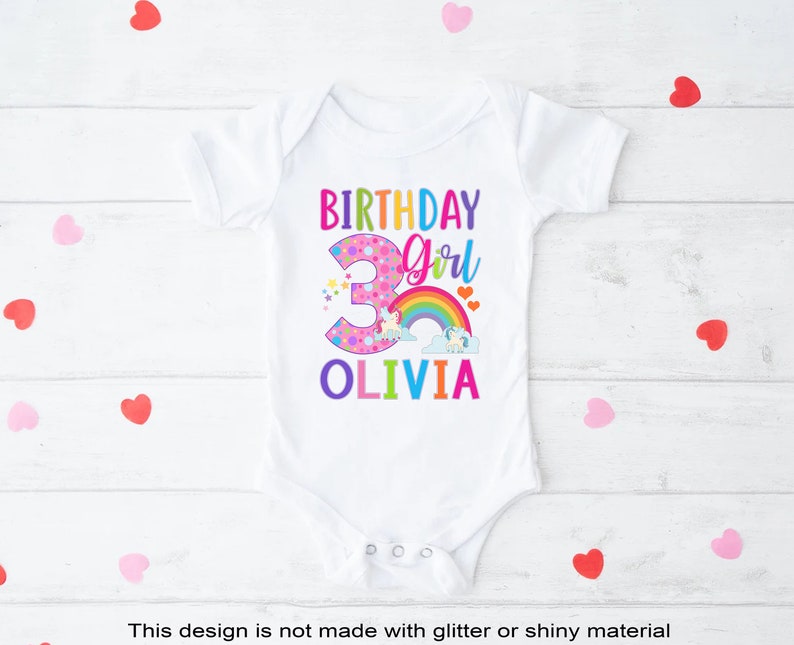 May include: White onesie with a colorful birthday girl design. The number 3 is pink with white polka dots and surrounded by stars. A rainbow with clouds and a unicorn is above the number. The text "Birthday Girl" is in rainbow colors and the name "Olivia" is in rainbow colors below the number.