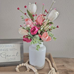 May include: A white ceramic vase filled with pink, white, and fuchsia tulips and greenery. The vase sits on a wooden tray with a beaded garland and a framed sign that reads "DO SMALL THINGS WITH GREAT LOVE."