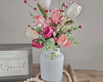 May include: A white ceramic vase filled with pink, white, and fuchsia tulips and greenery. The vase sits on a wooden tray with a beaded garland and a framed sign that reads "DO SMALL THINGS WITH GREAT LOVE."