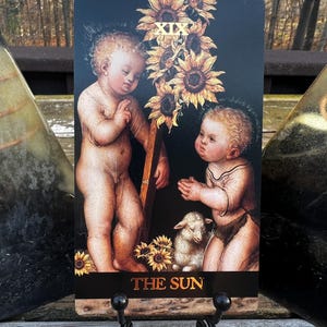 May include: A tarot card featuring two cherubic figures, sunflowers, and a lamb against a black background. The card is labeled "THE SUN" in gold lettering. The card is displayed on a black metal stand.