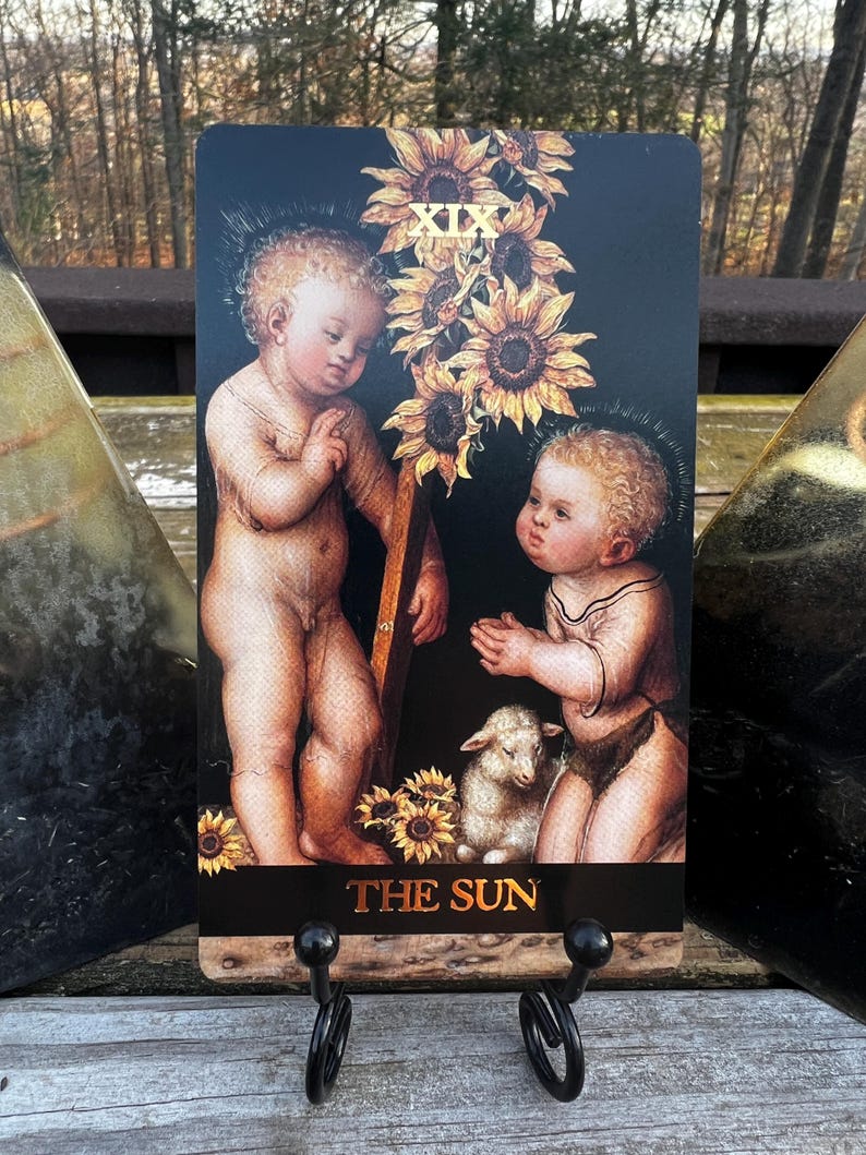 May include: A tarot card featuring two cherubic figures, sunflowers, and a lamb against a black background. The card is labeled "THE SUN" in gold lettering. The card is displayed on a black metal stand.