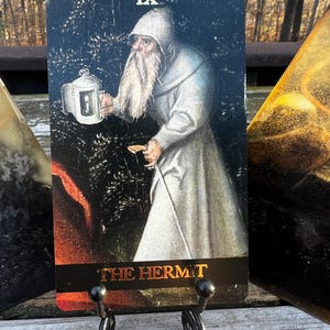 May include: A tarot card depicting "The Hermit" in a vintage style. The card shows a robed figure holding a lantern and staff, with the Roman numeral IX at the top. The card is displayed on a black metal stand.