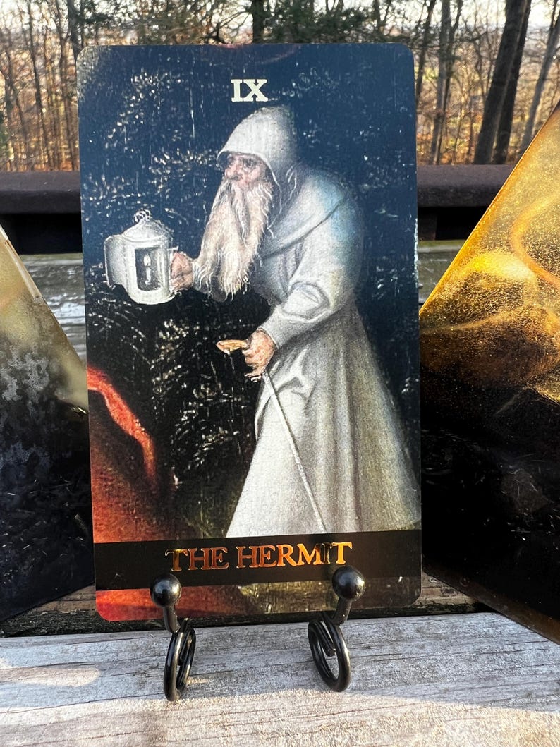 May include: A tarot card depicting "The Hermit" in a vintage style. The card shows a robed figure holding a lantern and staff, with the Roman numeral IX at the top. The card is displayed on a black metal stand.