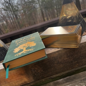 May include: A green Medieval Earth Tarot guidebook with gold lettering and a gold-edged deck of cards. The book and cards are on a wooden surface, with a clear pyramid-shaped object in the background.
