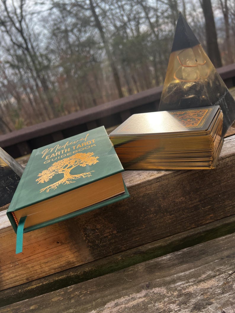 May include: A green Medieval Earth Tarot guidebook with gold lettering and a gold-edged deck of cards. The book and cards are on a wooden surface, with a clear pyramid-shaped object in the background.