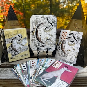 May include: A collection of Medieval Oracle of Lenormand cards. The cards feature illustrations with a vintage aesthetic, including a crescent moon design and the alphabet. The deck is displayed with two pyramid-shaped objects in the background.