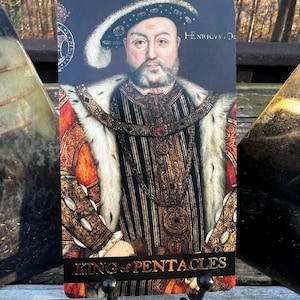 May include: A tarot card featuring a portrait of a man in regal attire, with a fur-lined robe and ornate jewelry. The card is labeled "King of Pentacles" in gold lettering. The background is dark, and the card is displayed on a black metal stand.