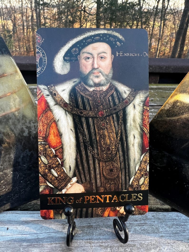 May include: A tarot card featuring a portrait of a man in regal attire, with a fur-lined robe and ornate jewelry. The card is labeled "King of Pentacles" in gold lettering. The background is dark, and the card is displayed on a black metal stand.