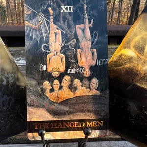 May include: Tarot card featuring an illustration of two figures hanging upside down, with a group of people below. The card is labeled "XII" and "THE HANGED MEN" in gold lettering. The card is displayed on a black metal stand.